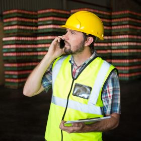 Male worker holding clipboard while talking on phone in factory