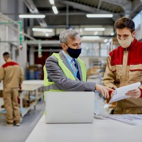 Factory worker and businessman with protective face masks analyzing reports at woodworking production facility.